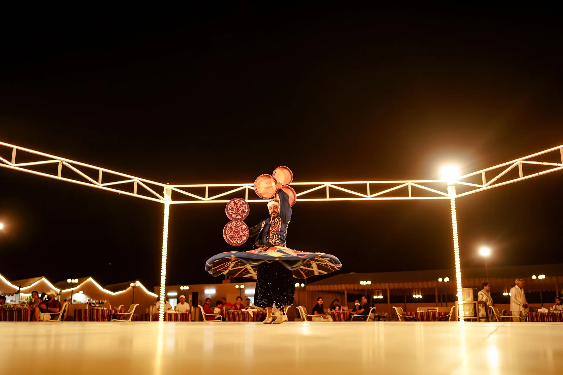 A Tanoura dancer spins under a lit steel arch on an open-air stage at a desert dinner show, dining tables and tented pavilions visible behind (representative photograph of a Tanoura performance in the UAE desert, not Al Hadheerah's own stage)